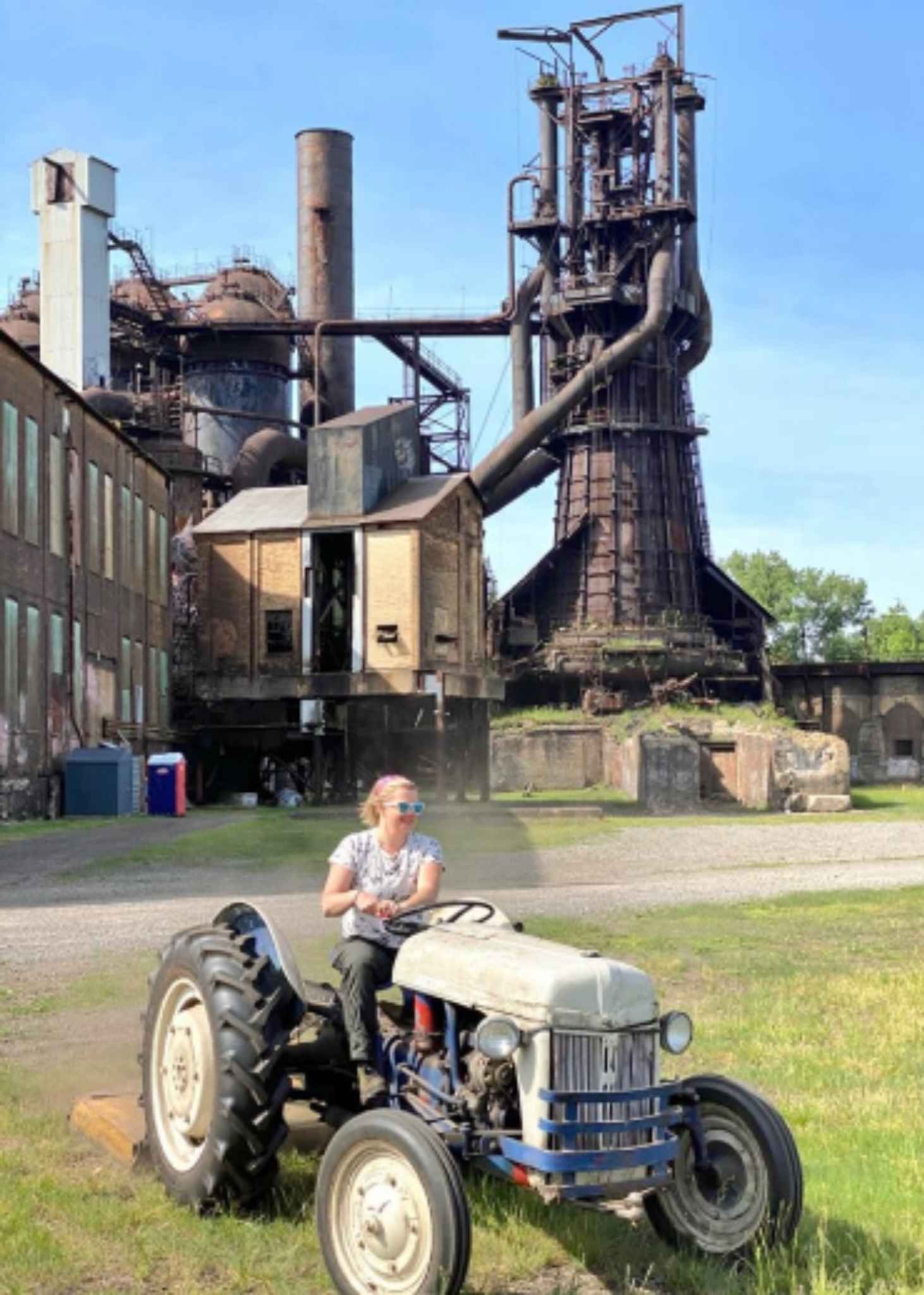 Alexandra Hiniker driving a vintage tractor across a lawn in front of the rusted towers of the Carrie Blast Furnaces