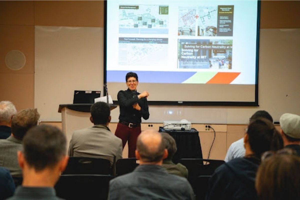 Julie Newman presenting to a seated audience in a lecture hall, with photos of campus projects on the screen behind her