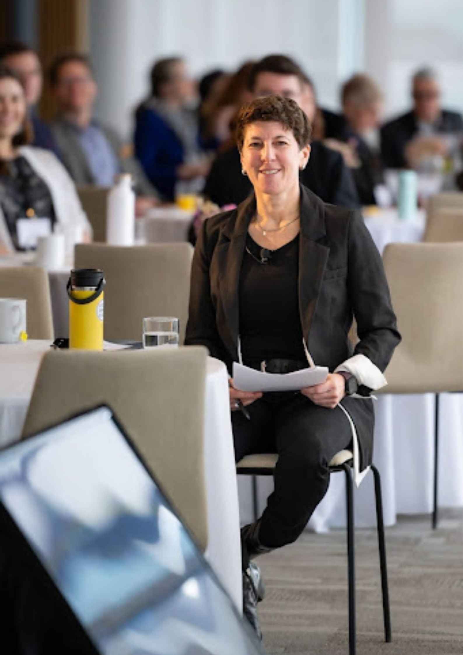 Julie Newman seated in the front row of a conference audience, holding notes and smiling