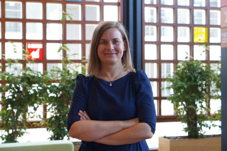 Portrait of Dr. Simone Sandholz of UNU-EHS, smiling with arms folded in front of a large window and plants