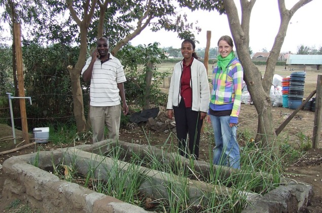 Three diverse community members standing proudly next to a raised vegetable garden bed in a rural setting, showcasing a local, sustainable agriculture project.