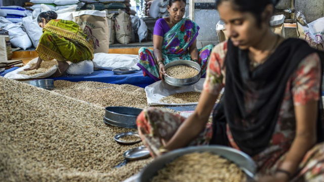 Three women in a rural market manually cleaning and sifting harvested grain with traditional sieves, a crucial step in post-harvest processing.