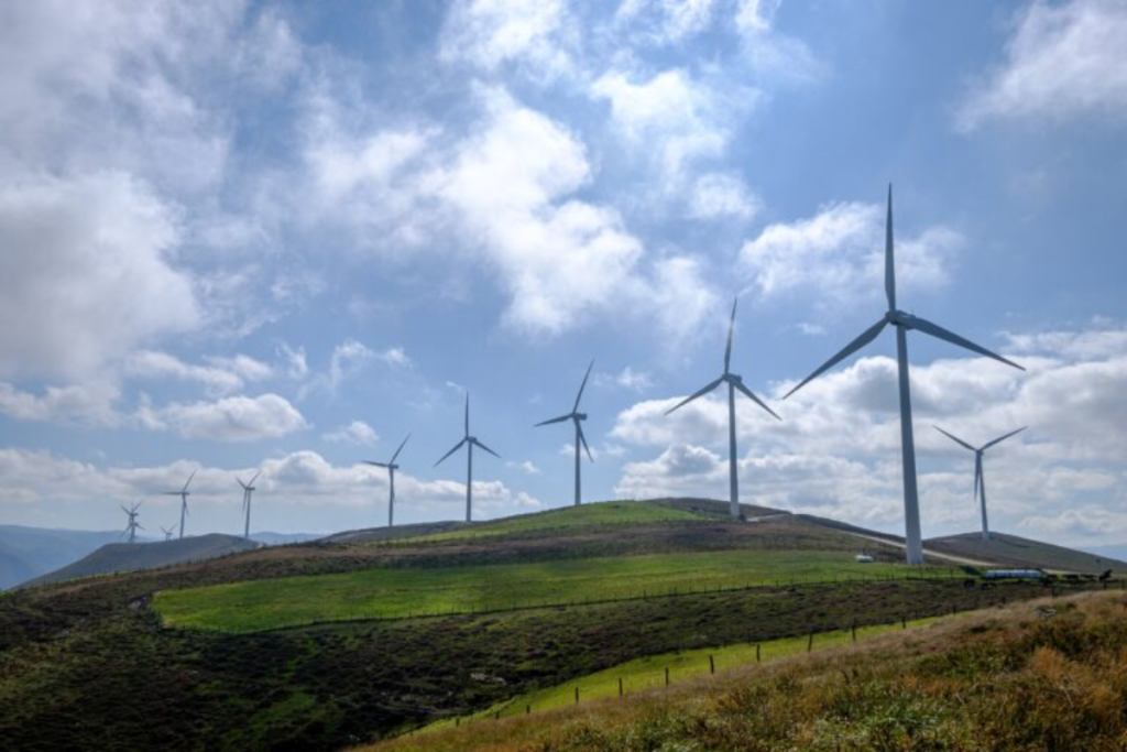 A row of modern wind turbines on a lush green hillside under a partly cloudy sky, representing renewable energy and environmental sustainability.