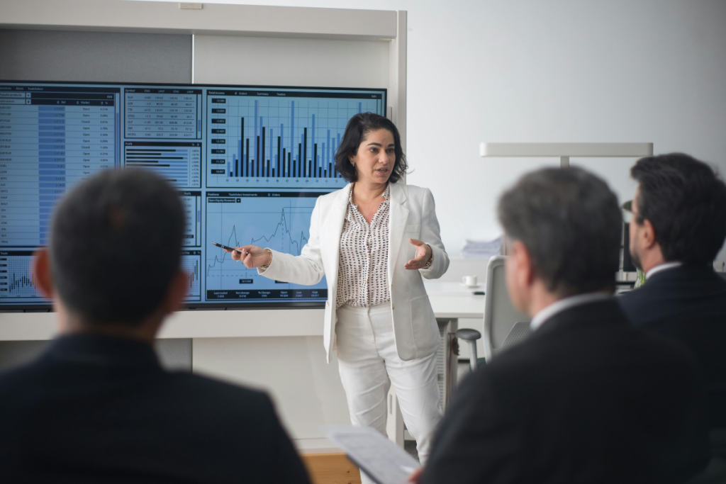 A female executive in a white suit presenting complex financial and ESG data charts on large screens to a group of business professionals.