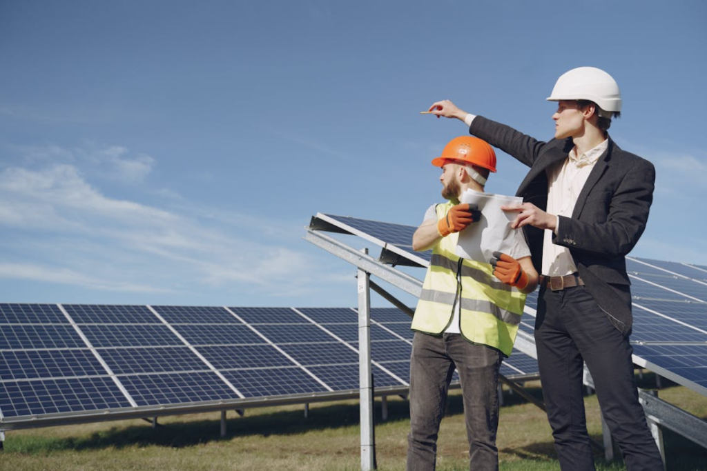 An engineer in a suit and a worker in a safety vest stand together at a solar farm, discussing plans and pointing towards the solar panels.
