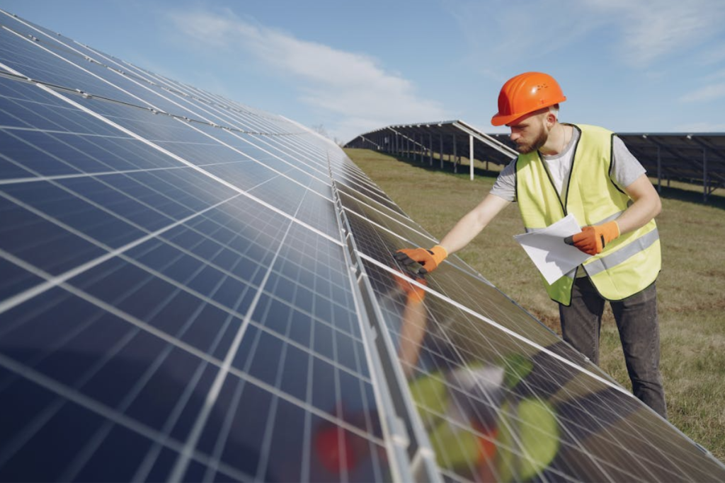 An engineer in an orange hard hat and yellow safety vest inspects a solar panel in a large solar farm on a sunny day.