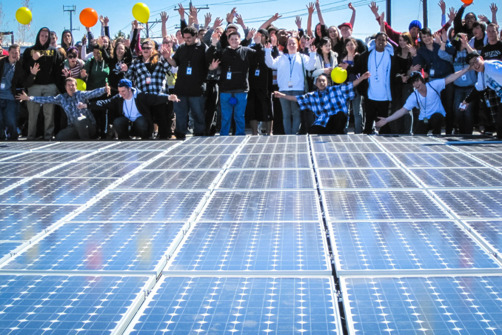 A large, diverse group of enthusiastic people celebrating with their hands in the air behind a large installation of solar panels.