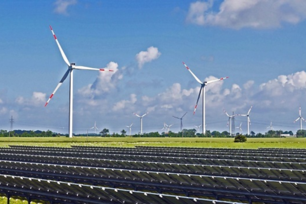 A hybrid renewable energy farm with rows of solar panels in the foreground and large wind turbines spinning in the background under a clear blue sky.
