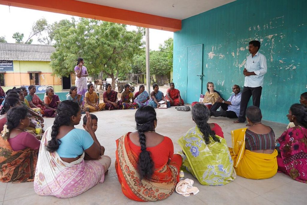 A community meeting in a rural Indian village, where a Women's Self-Help Group (SHG) sits in a circle, listening to a trainer from Earth5R
