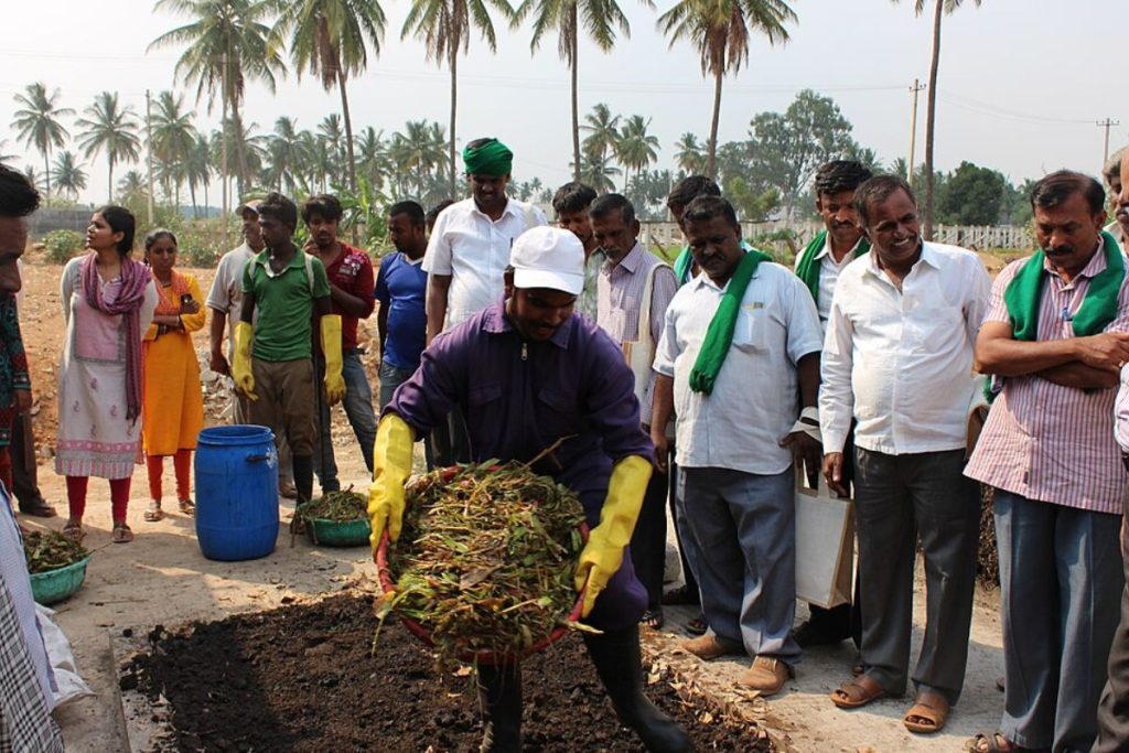 An Earth5R trainer in protective gear demonstrates composting techniques to a group of Indian farmers, showcasing sustainable waste management