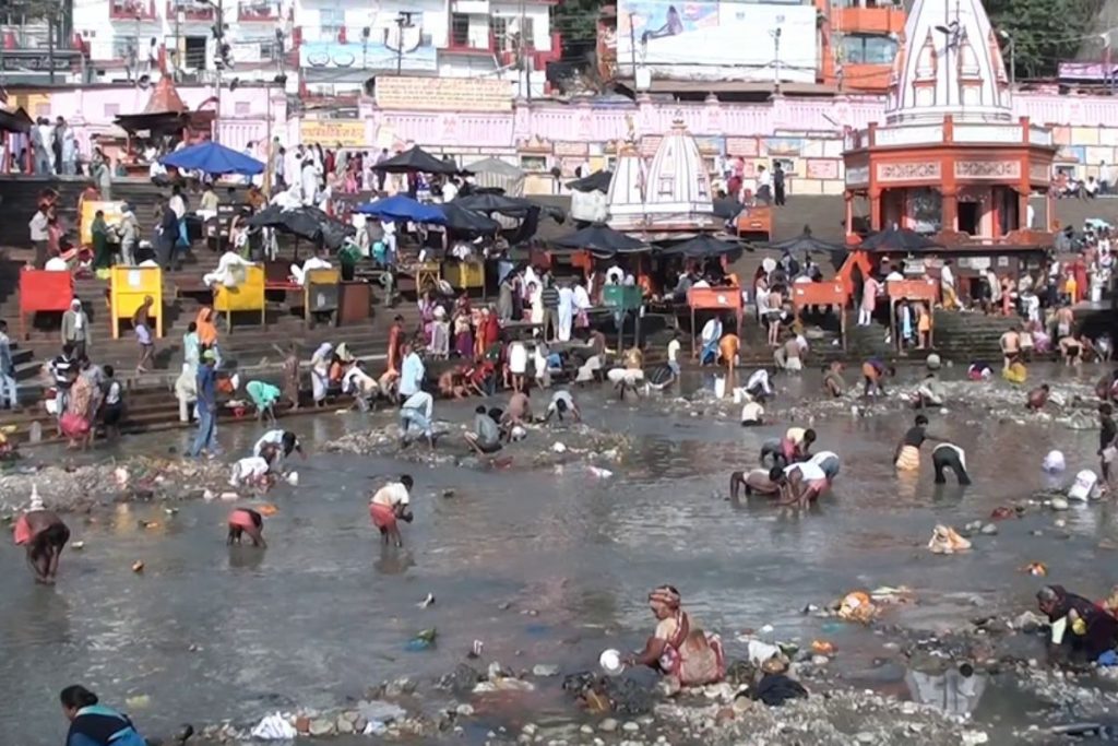 A crowded pilgrimage site on an Indian river, filled with people bathing, while the water and banks are visibly littered with plastic bags, bottles, and other trash.