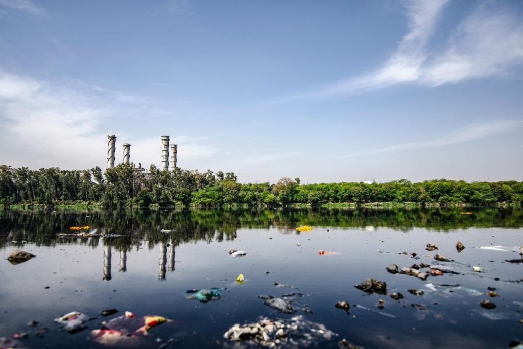 A dark, polluted river with floating plastic trash in the foreground and a large industrial plant with chimneys reflected in the water on the far bank.