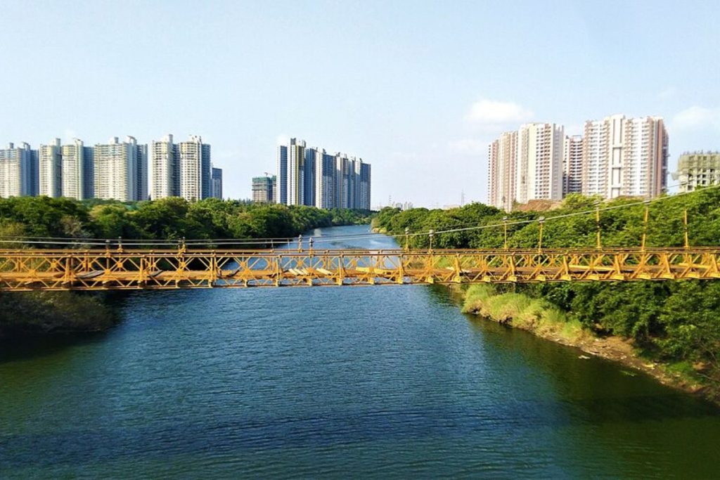 A wide urban river with clean, blue water flowing under a yellow bridge, flanked by green trees and modern high-rise apartments.