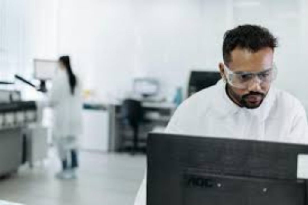 Scientist in a lab coat and safety goggles working at a computer, focused on environmental data analysis.