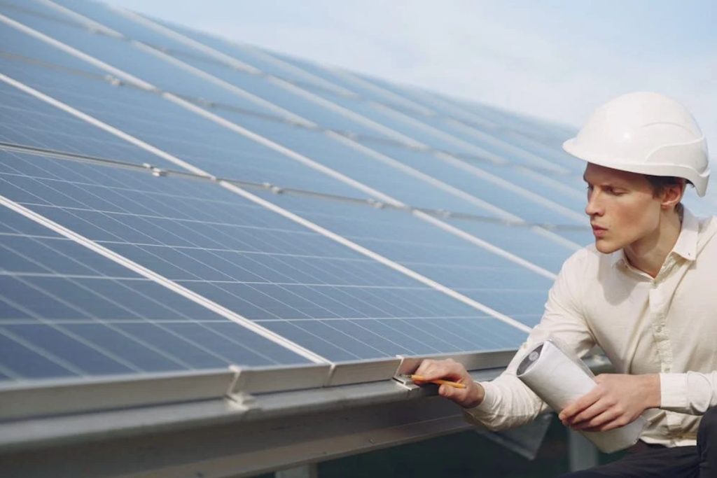 An engineer in a white hard hat inspects a solar panel array up close, holding a clipboard and pencil.