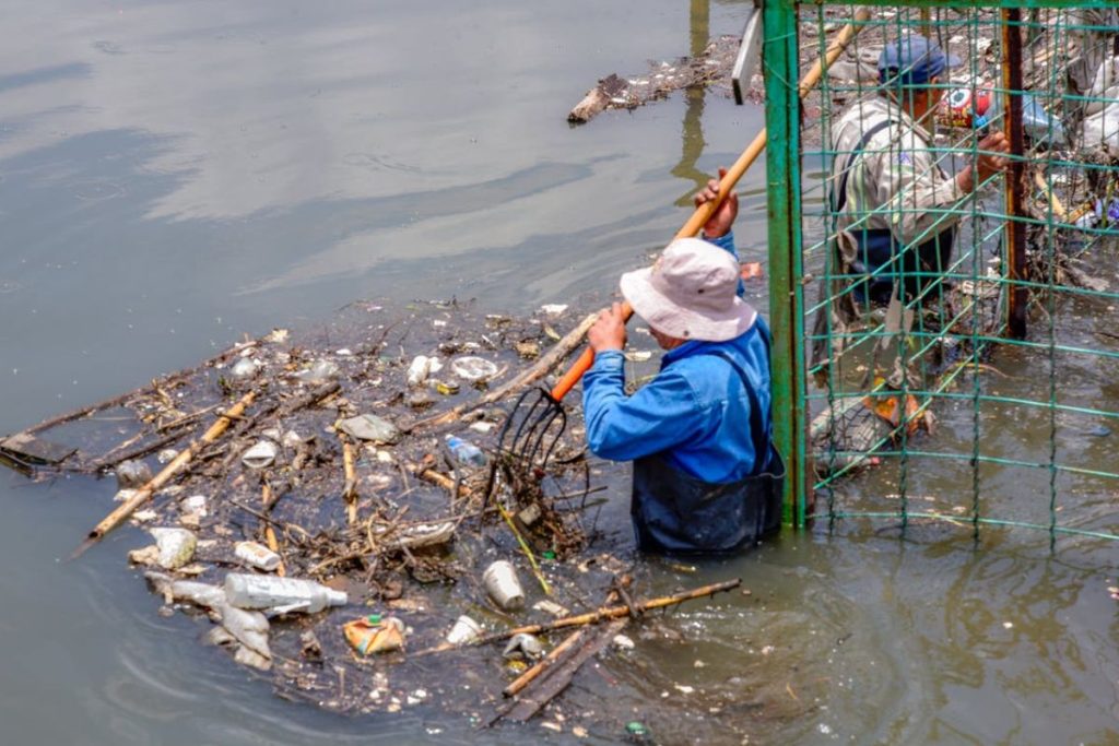 Workers stand waist-deep in a polluted Mumbai river, sorting through piles of garbage as part of a crucial waste management effort, with a crowded urban settlement in the background.
