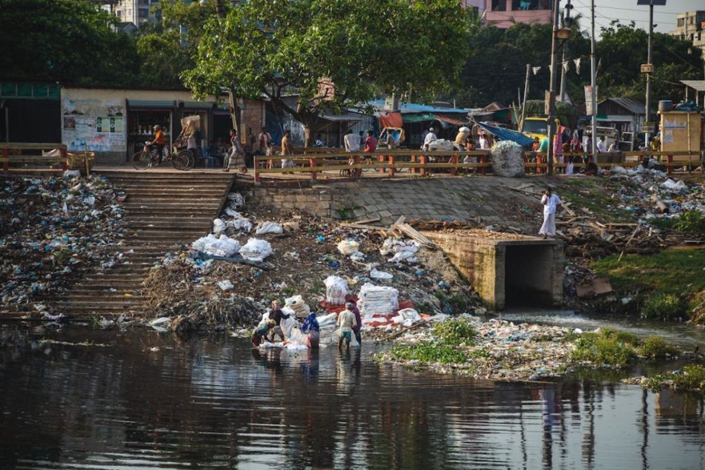 A close-up of two workers in waders using rakes to collect a dense patch of plastic bottles and other floating debris from a river, showcasing a hands-on waste management initiative.
