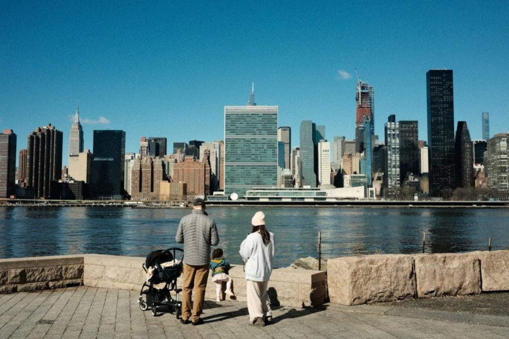 A family with a young child and stroller enjoys a walk along a clean, wide river, looking at the New York City skyline, representing a future of urban sustainability.