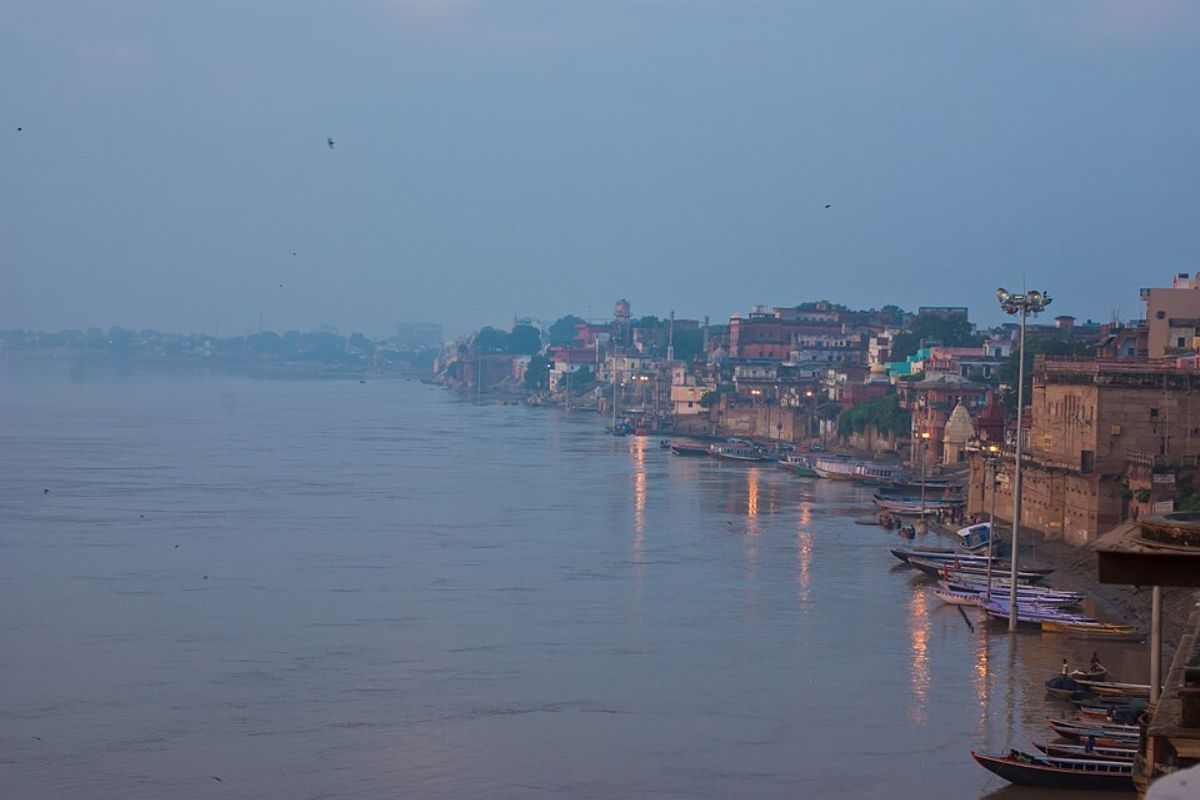 A wide view of the Ganga river at dusk, showing the historic ghats of Varanasi and boats docked along the bank.