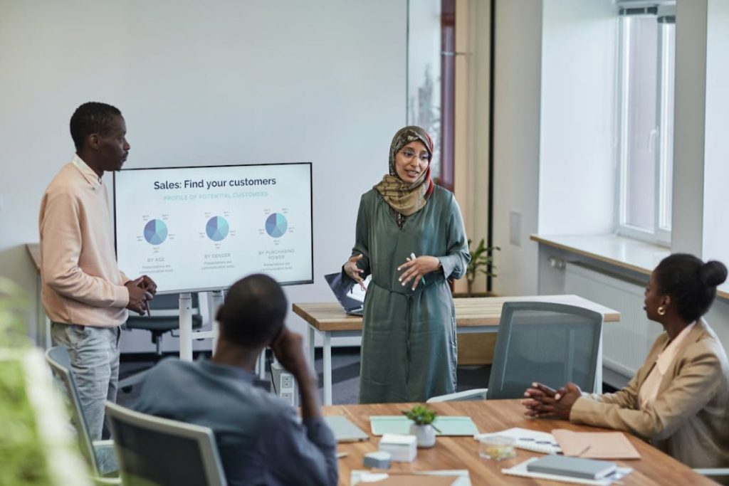 A woman in a hijab confidently presents data to a diverse team in a modern office, illustrating an employee-driven sustainability initiative and a culture of empowerment.