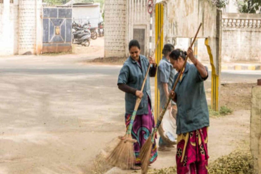 Women sanitation workers sweeping a roadside area, maintaining cleanliness as part of local community waste management efforts.