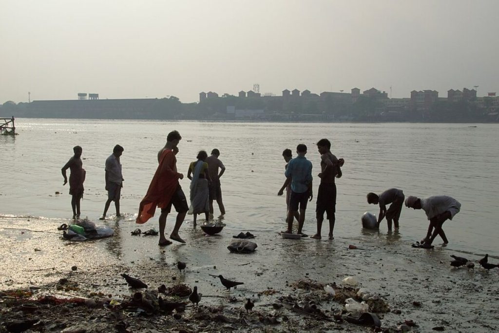 People wash clothes and bathe on the polluted, trash-strewn bank of a river, illustrating the direct human impact of water pollution.