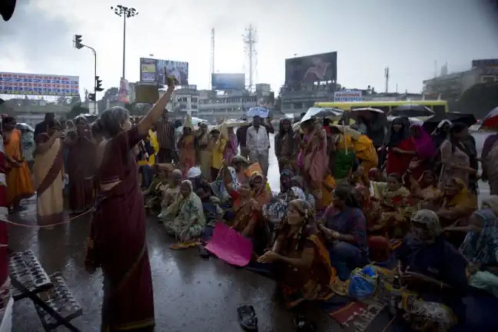 A large group of community members, mostly women, sit on a rain-soaked city street during a gathering or protest.