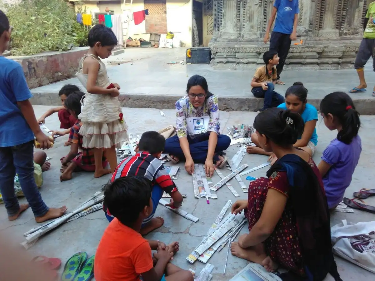 Earth5R volunteer and children making newspaper craft sitting on the ground outdoors