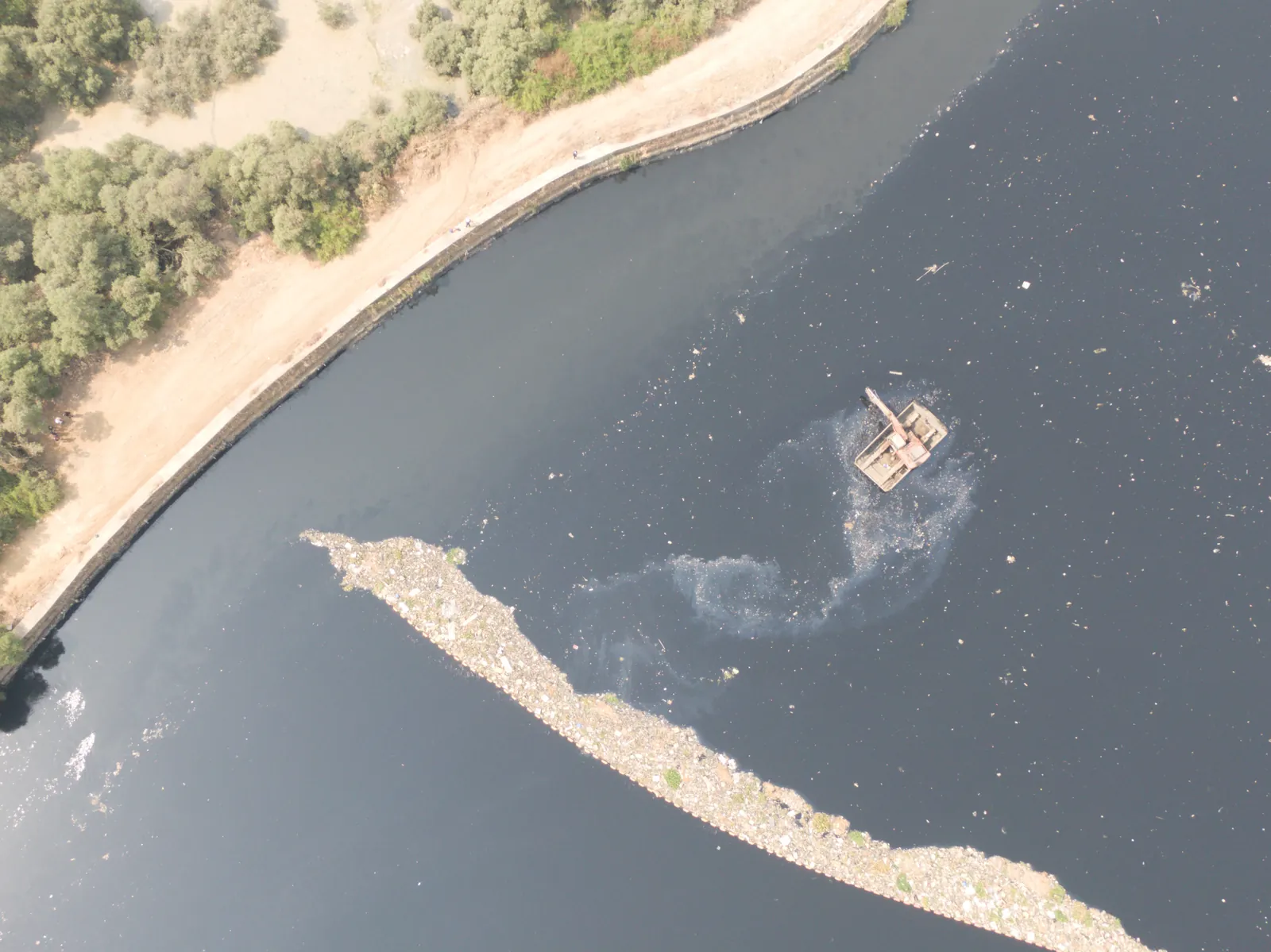 GIS internship field work. Aerial view of a dark river with a line of floating waste held along the bank