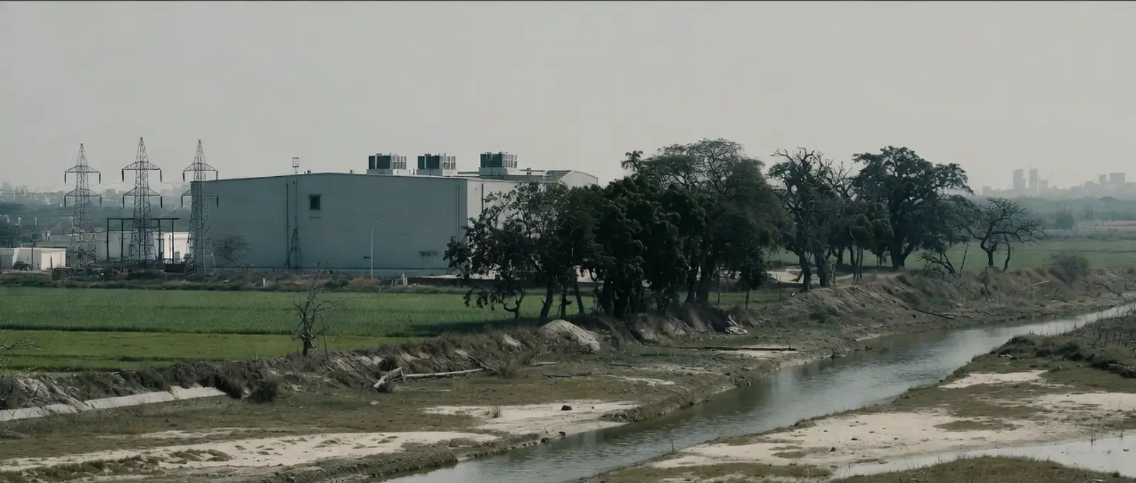 A data centre with rooftop cooling units beside farmland, trees and a low river on the edge of an Indian city