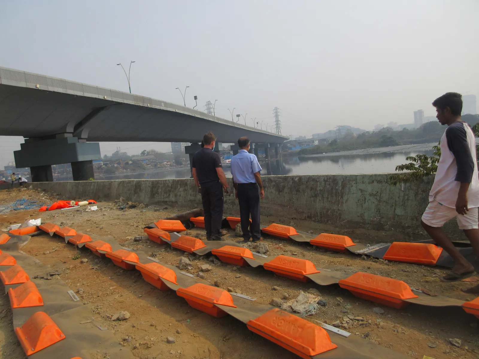 Volunteers assembling orange trash boom floats on a riverbank below a flyover