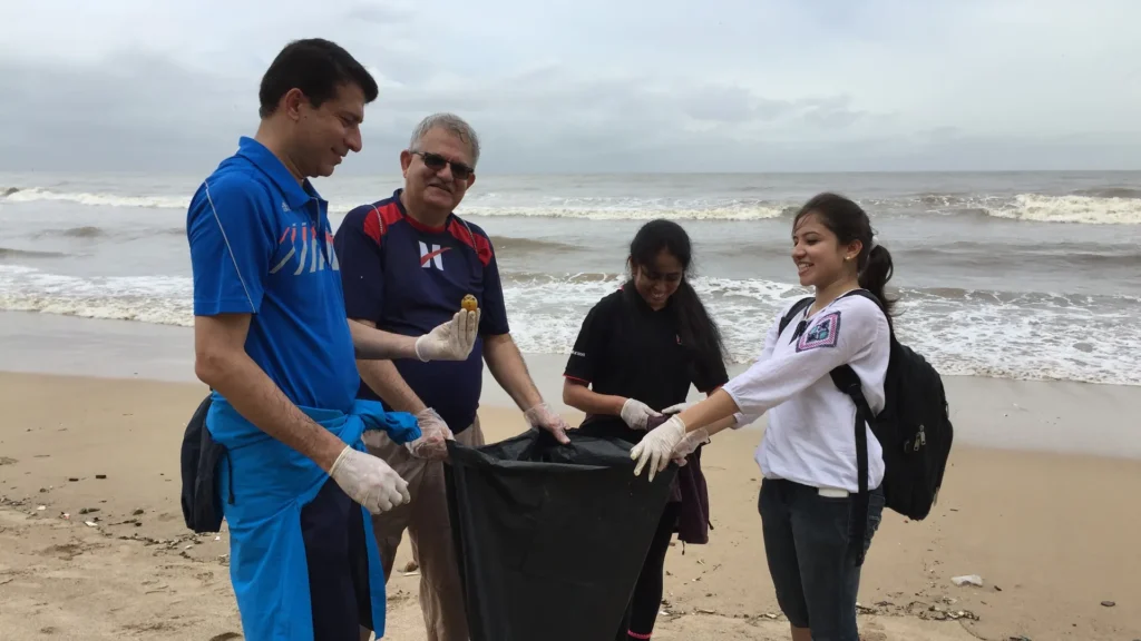 Four volunteers in gloves holding open a black garbage bag on a Mumbai beach during an Earth5R and Hexaware cleanup on International Coastal Cleanup Day, rough sea behind
