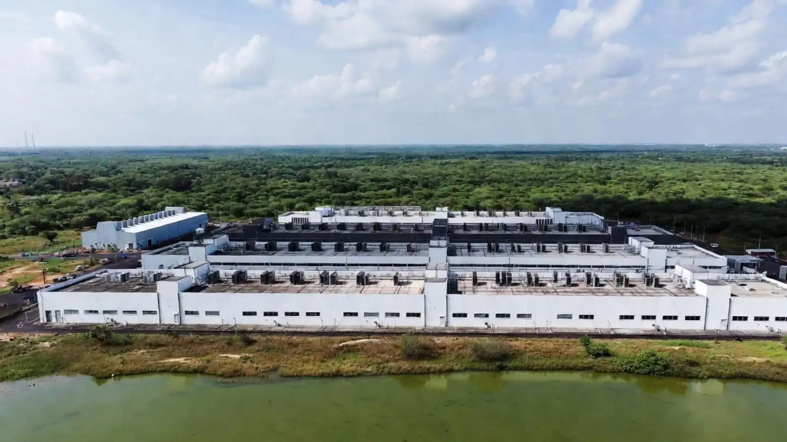 Long white data centre building with rooftop cooling units beside an urban lake with green algae, showing pressure on local water bodies
