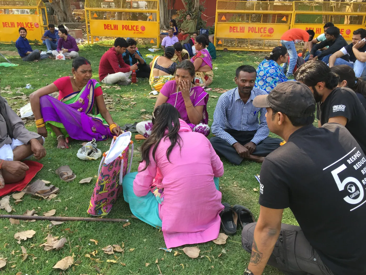 An Earth5R volunteer in a black T-shirt seated on the grass with women and men in a community training session, yellow barricades behind them