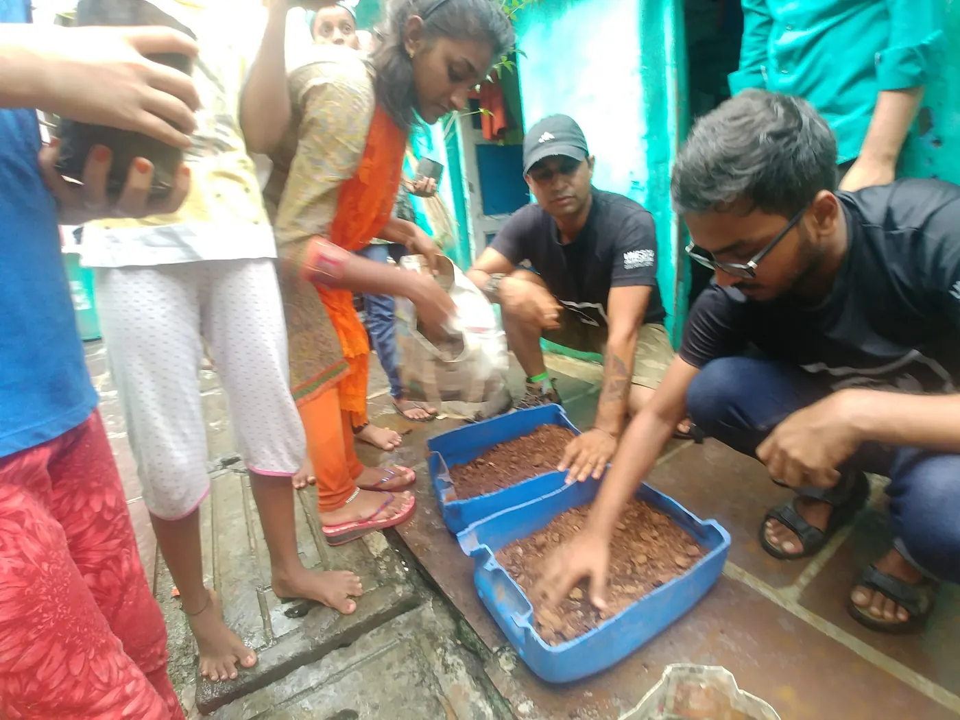 Earth5R volunteers and residents filling blue crates with compost and soil in a narrow lane of a Mumbai informal settlement, ready for planting saplings