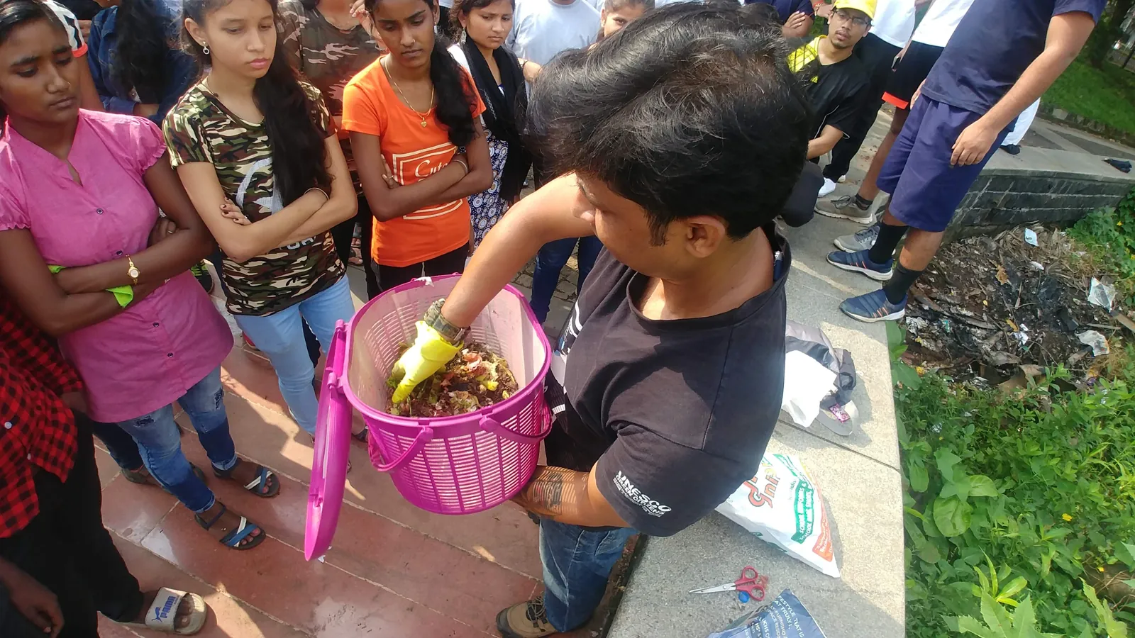 An Earth5R volunteer showing a pink compost basket to a crowd of young women and residents gathered on a lakeside path