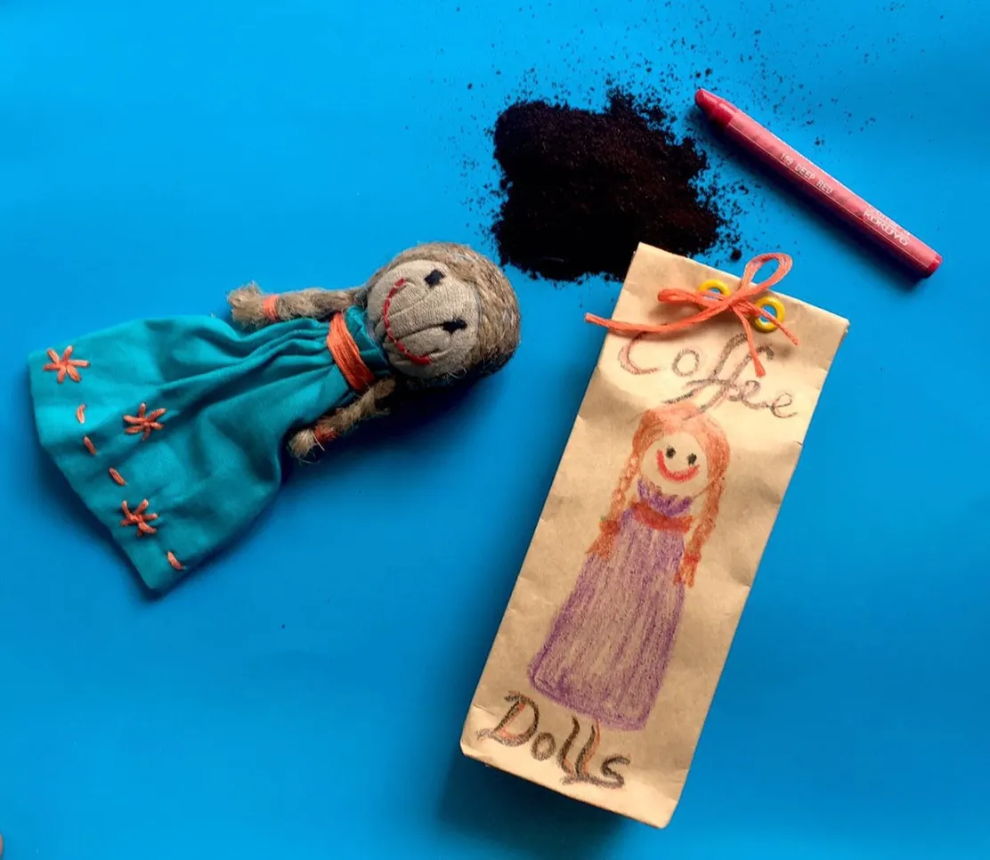 A small handmade doll in a blue dress stitched from fabric scraps, beside a paper tag reading Coffee Dolls, on a blue background