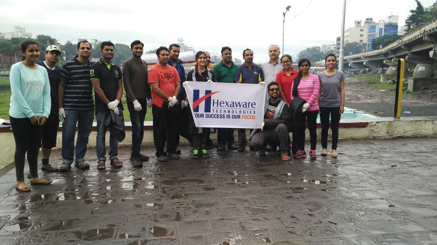 About fifteen Hexaware Technologies employees and Earth5R volunteers in gloves holding a Hexaware banner on a rain-wet seafront promenade before a beach cleanup, 2016