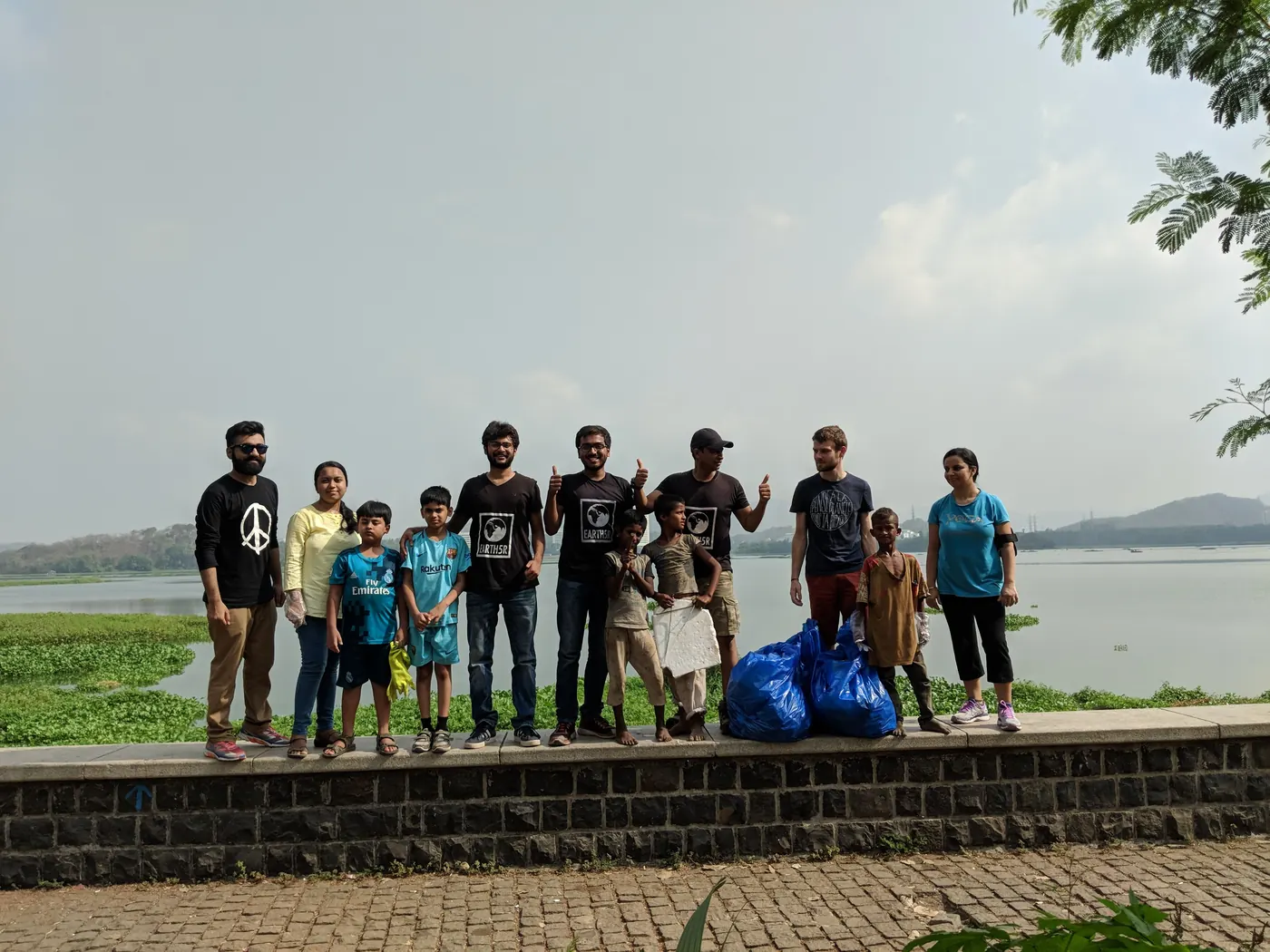 Earth5R volunteers and children standing on a lakeside wall with blue bags of collected waste, lake water behind them, 2018