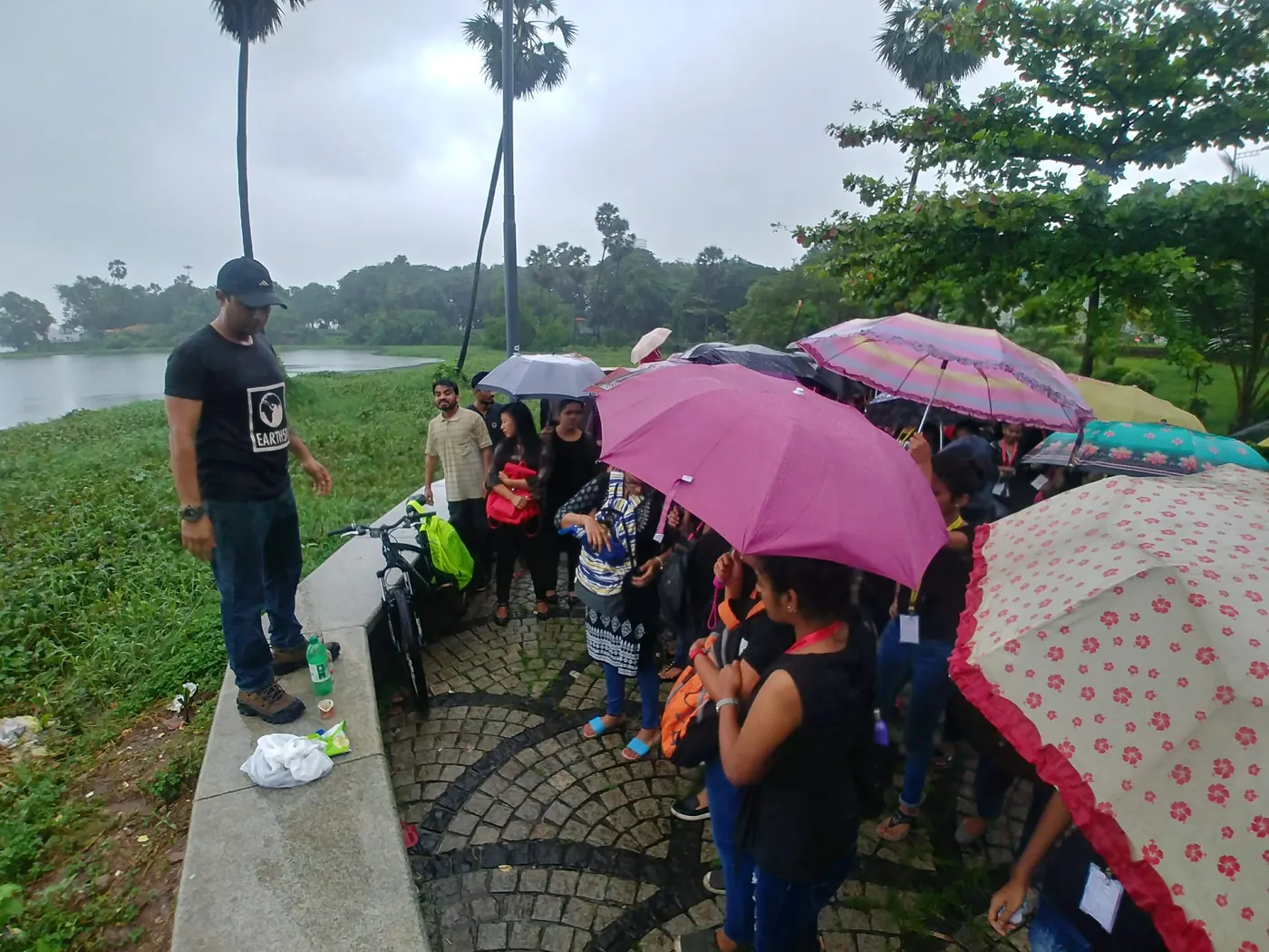 Earth5R volunteer briefing a crowd of young volunteers holding umbrellas on a lakeside path in monsoon rain, 2018