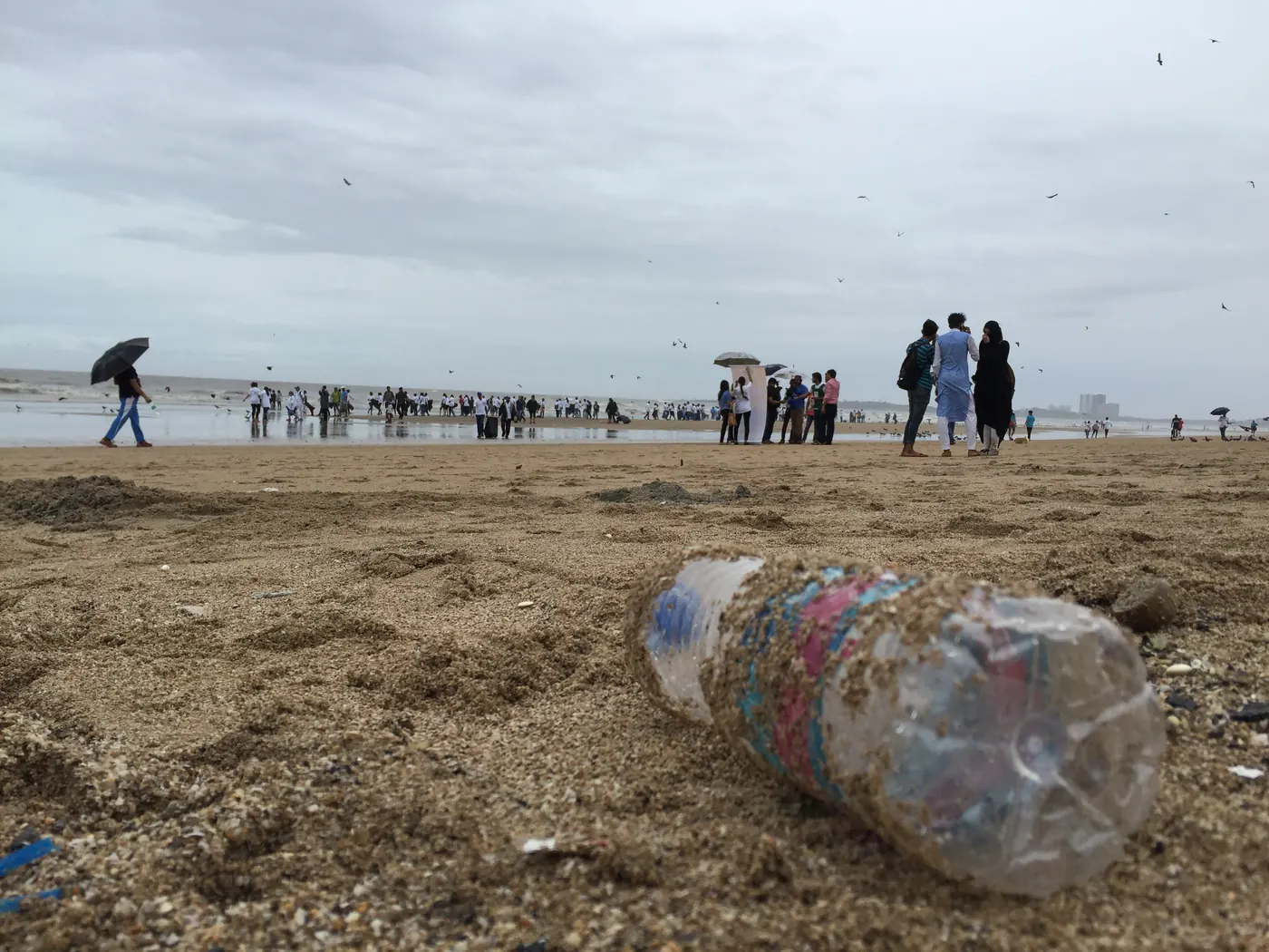 Crushed plastic water bottles lying on wet sand in the foreground of a Mumbai beach, with morning walkers along the shoreline behind