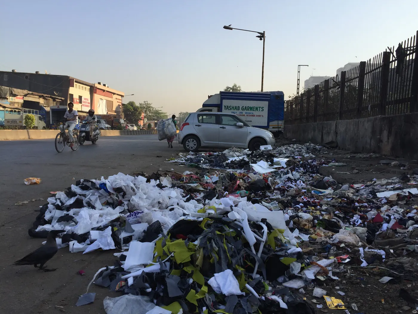 A long spread of plastic bags, wrappers and mixed garbage dumped along a shoreline road at dawn, with parked vehicles and a wall beside it
