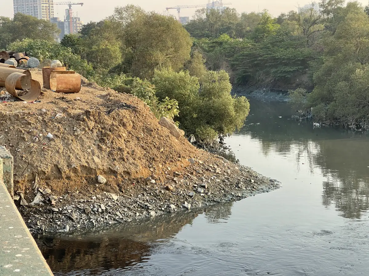A narrow urban creek with dark, still water, its bank piled with dumped debris and mixed waste, trees and buildings on either side