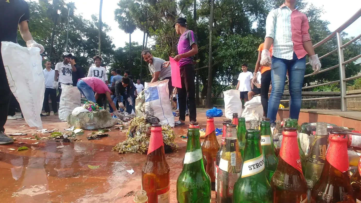 Rows of green and brown glass bottles lined up on wet tiles after a lake cleanup, volunteers with white sacks of sorted waste behind them