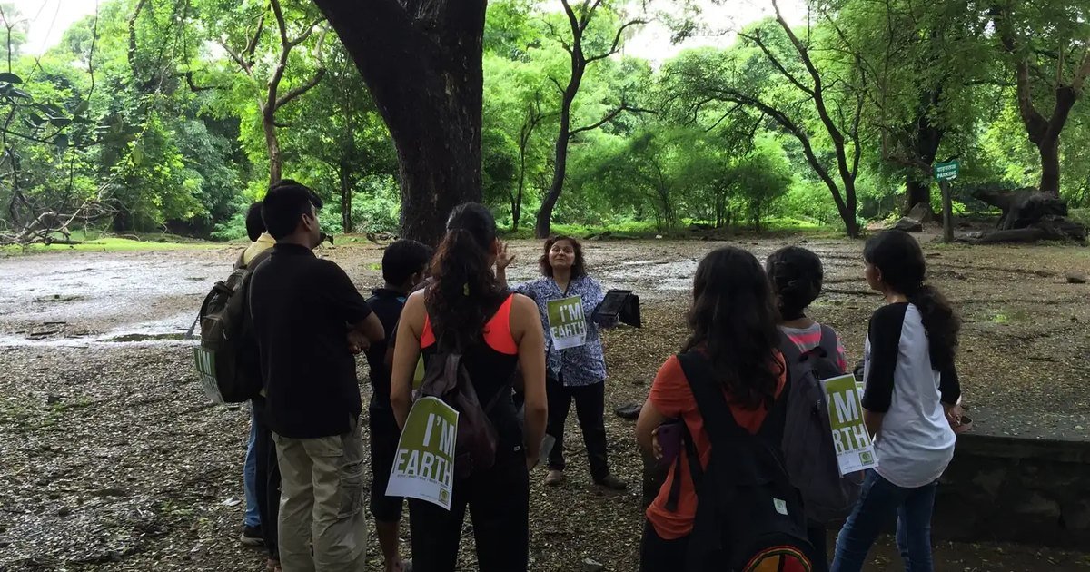 Students carrying Earth5R I am Earth signs listen to a guide under large trees on a forest nature trail in Mumbai