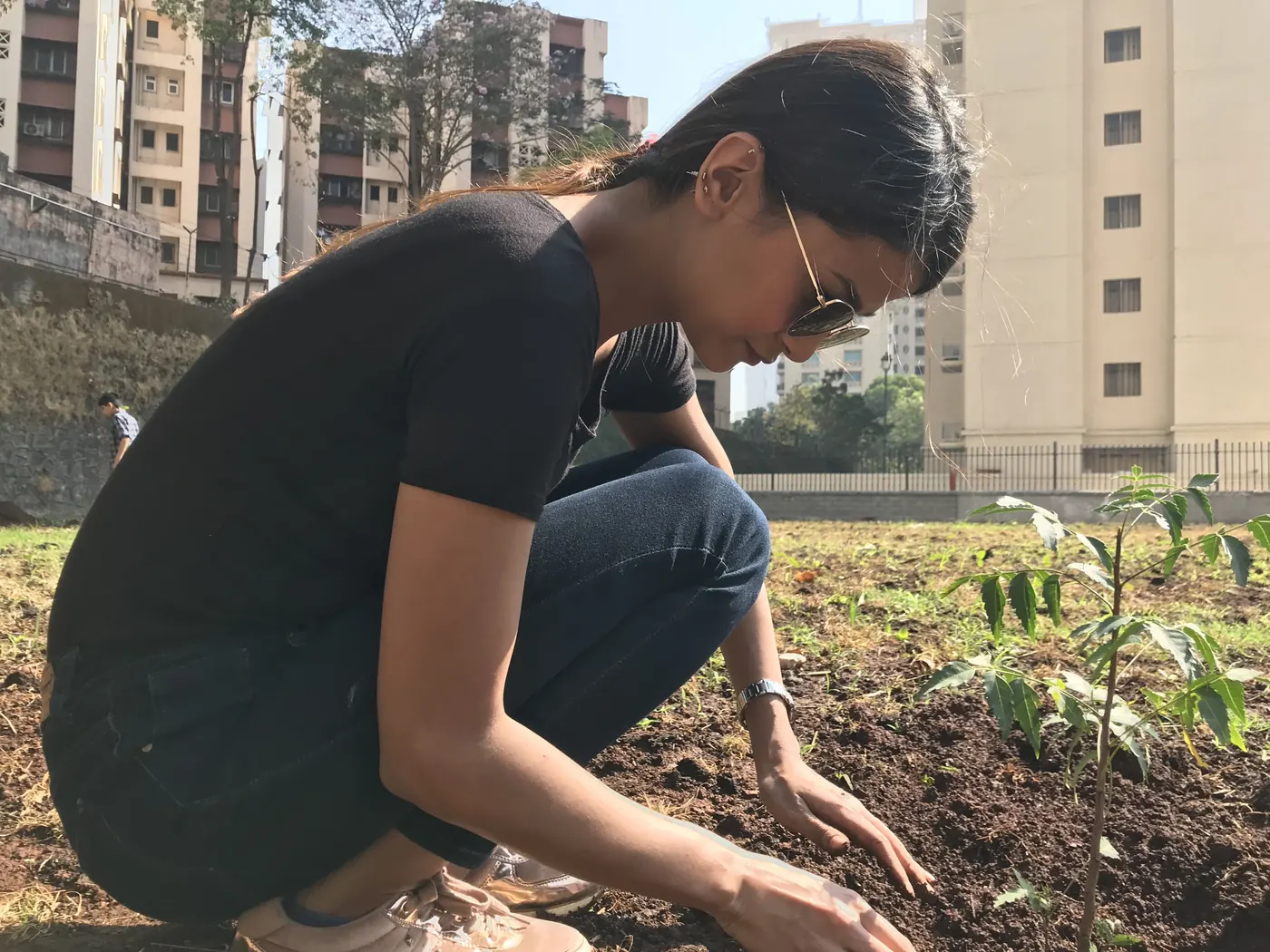 An Earth5R volunteer kneeling to plant a sapling in freshly dug red soil on an urban plot, with apartment towers behind
