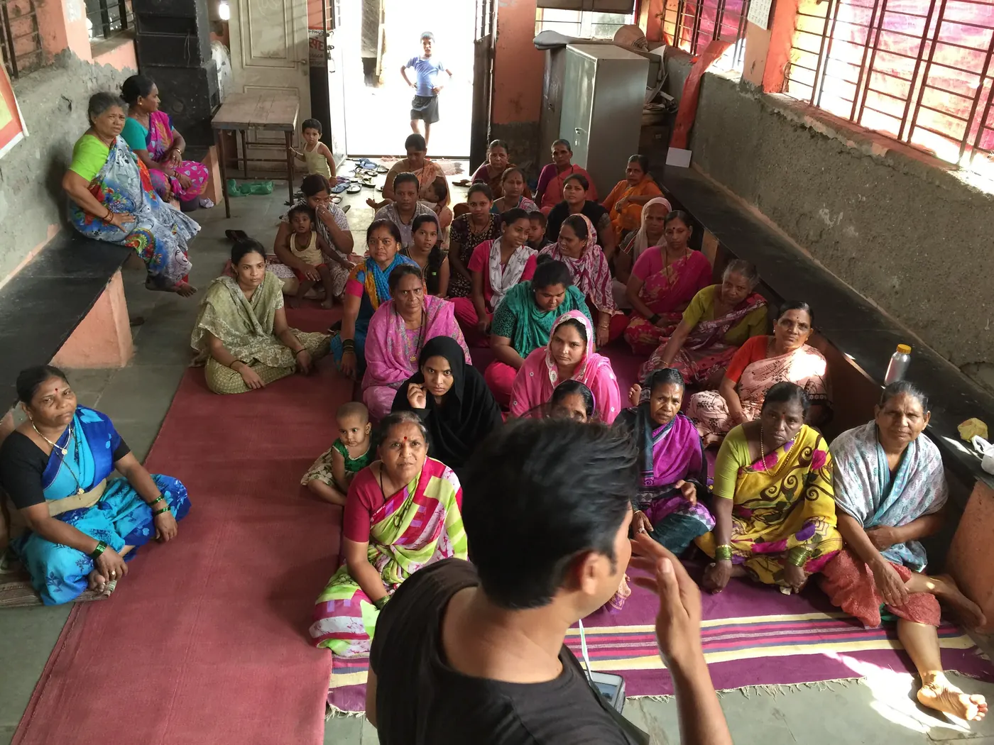 An Earth5R volunteer, seen from behind, speaking to a hall full of seated women at a community meeting