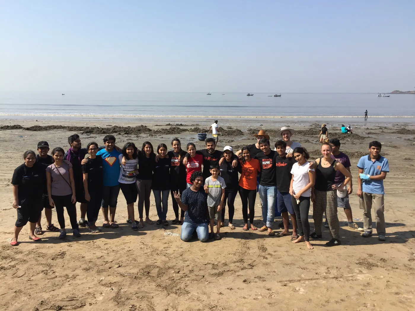 About forty Earth5R volunteers of all ages standing together on a wide sandy beach at low tide after a cleanup, fishing boats on the calm sea behind