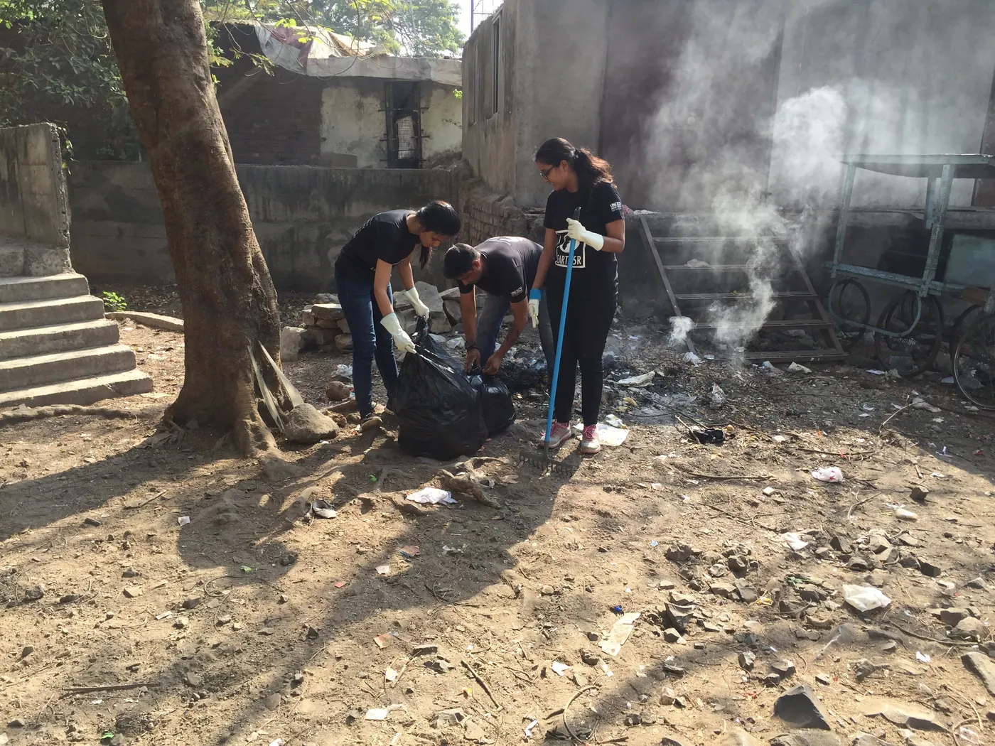 Earth5R volunteers in black T-shirts bagging waste in a dusty yard while white smoke rises from smouldering rubbish next to a building