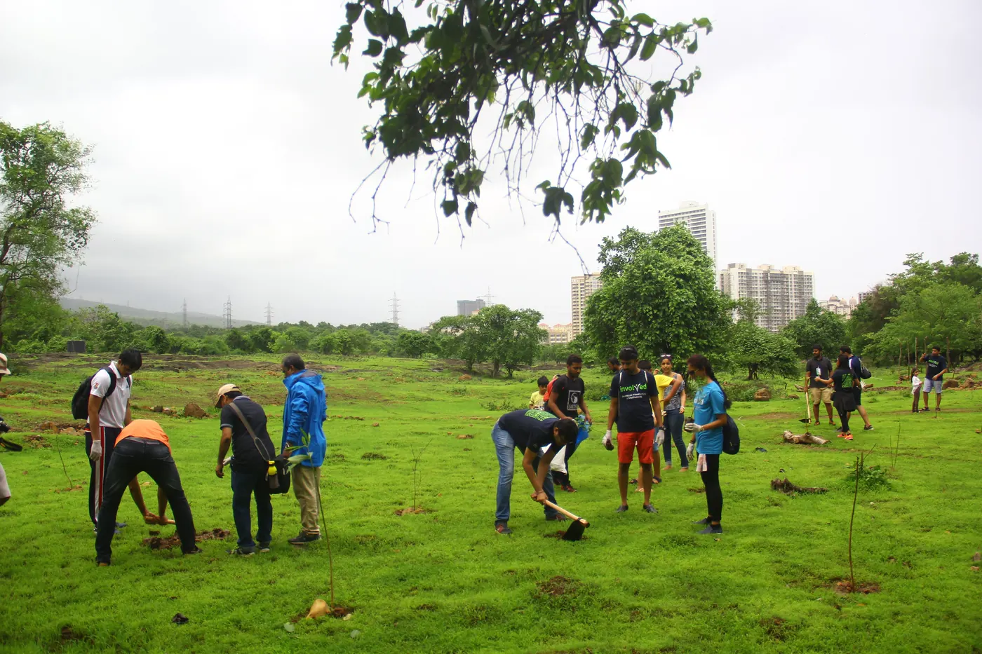 Dozens of Earth5R volunteers spread across a green field planting saplings under a cloudy monsoon sky, with trees and apartment towers on the skyline